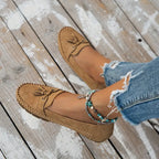 Mocassins en daim beige sur pieds féminins, associés à un jean déchiré et des bracelets de cheville turquoise, posés sur un sol en bois.