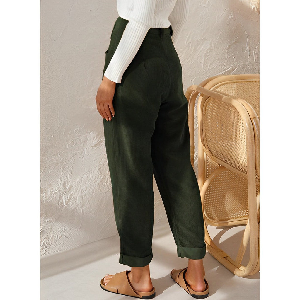 Femme portant un pantalon vert olive ample, associé à un pull blanc côtelé et des sandales marron, posant près d'une chaise en rotin.