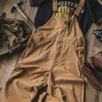 Salopette marron en toile robuste posée sur un sol en bois, accompagnée de gants de travail, sac à dos, marteau et bottes en cuir. Tenue de travail.