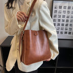 Femme en blazer blanc tenant un sac à main en cuir marron élégant, moderne et tendance, devant une étagère noire et un cadre photo.