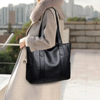 Femme portant un sac à main en cuir noir élégant, manteau beige avec col en fourrure, vue urbaine en arrière-plan. Mode chic et moderne.