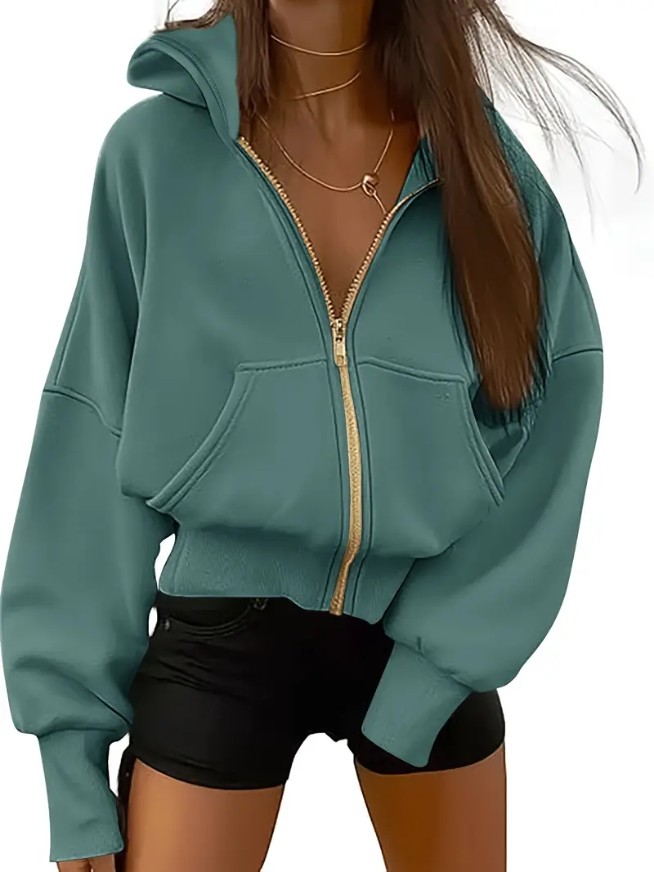 Femme portant un sweat à capuche vert avec fermeture éclair dorée, manches longues, poches avant, collier doré, et short noir. Mode décontractée tendance.