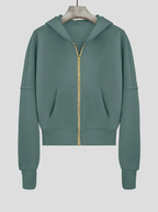 Sweat à capuche vert avec fermeture éclair dorée, manches longues, poches kangourou, suspendu sur cintre. Mode décontractée, vêtement tendance.