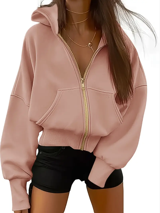 Femme portant un sweat à capuche rose avec fermeture éclair dorée, colliers superposés, et short noir. Mode décontractée, tendance automne.