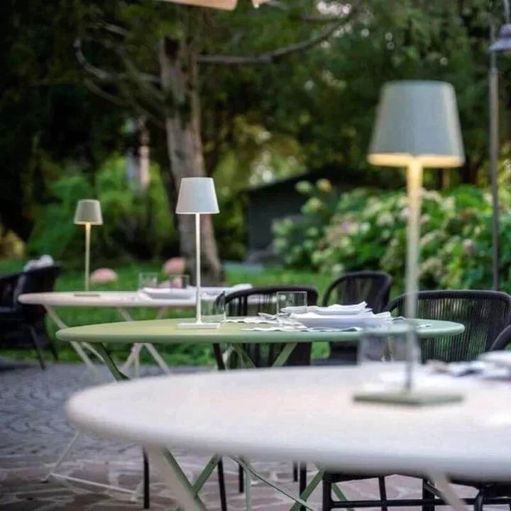 Tables de jardin élégantes avec lampes modernes, nappes blanches et chaises noires, situées dans un cadre verdoyant et paisible.