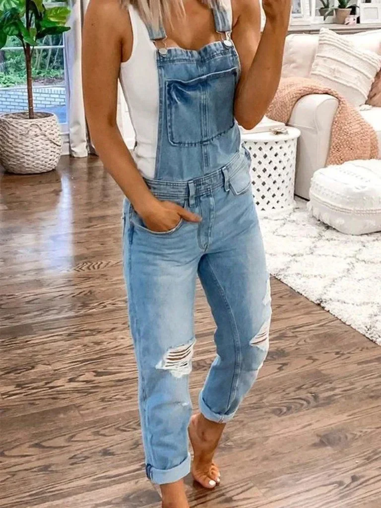Femme portant une salopette en jean déchiré, posant dans un salon moderne avec parquet, canapé blanc et plantes d'intérieur. Mode décontractée tendance.