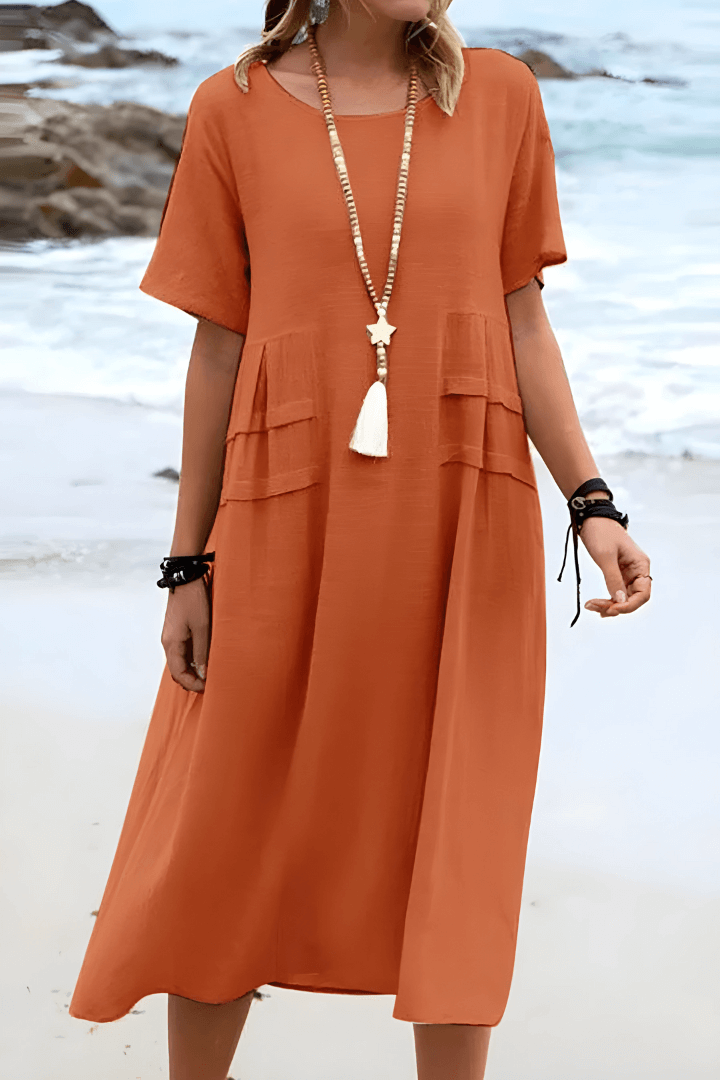 Robe orange en lin, manches courtes, portée par une femme sur une plage. Style décontracté, collier long, ambiance estivale.
