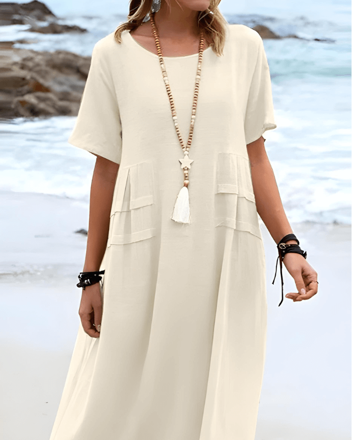 Robe longue beige en lin, manches courtes, col rond, avec collier à pompon blanc, portée par une femme sur une plage rocheuse en bord de mer.