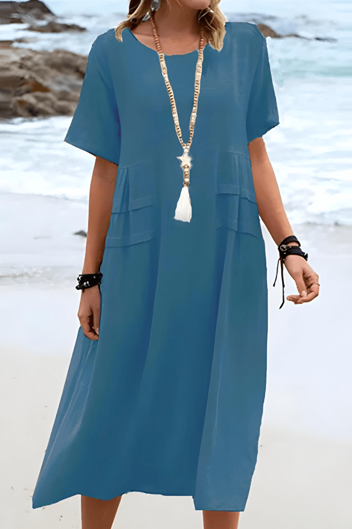 Robe bleue d'été fluide portée par une femme sur la plage, accessoirisée d'un long collier à pompon blanc. Mode plage élégante et décontractée.