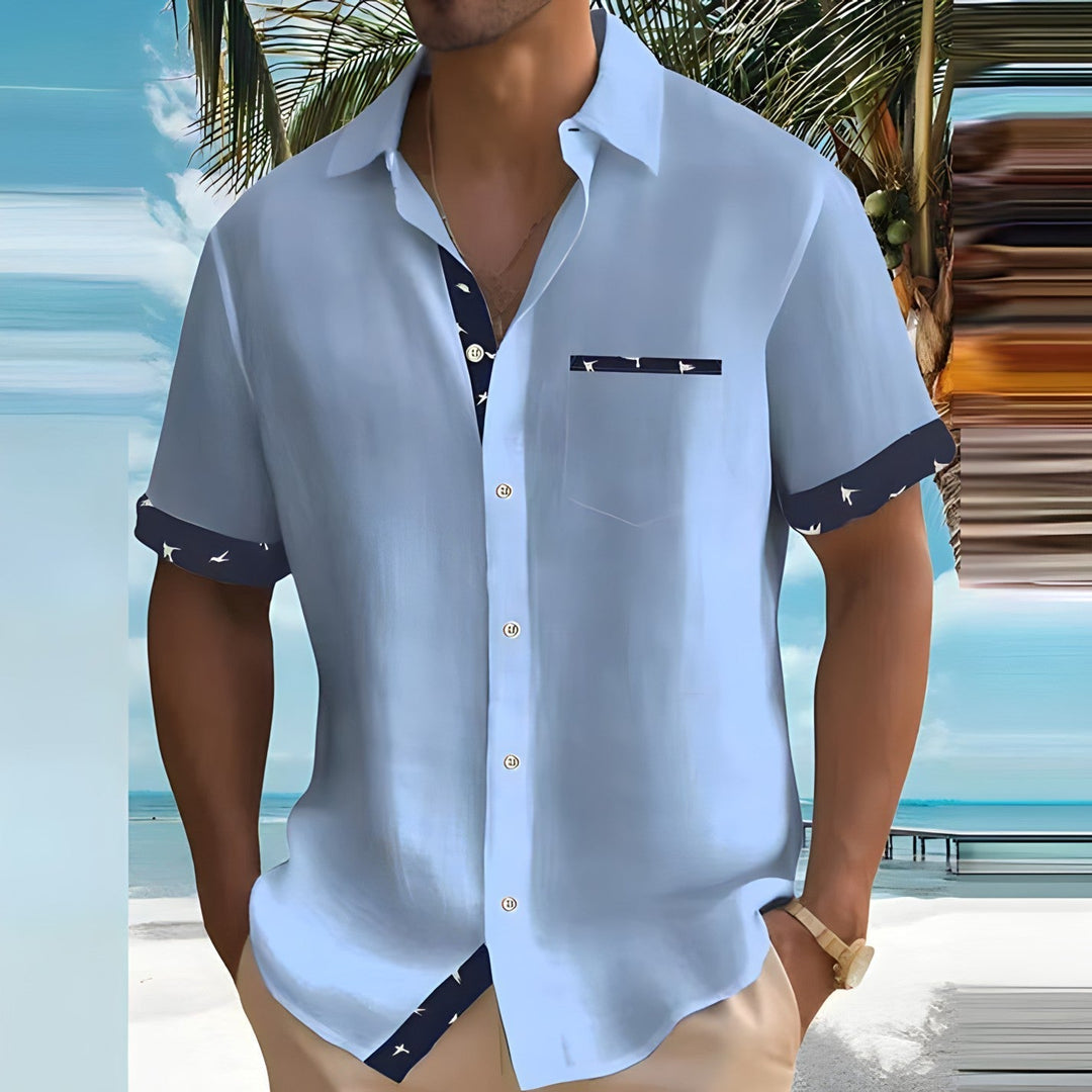 Homme en chemise bleue à manches courtes sur plage tropicale, palmiers en arrière-plan, style estival décontracté, mode vacances.
