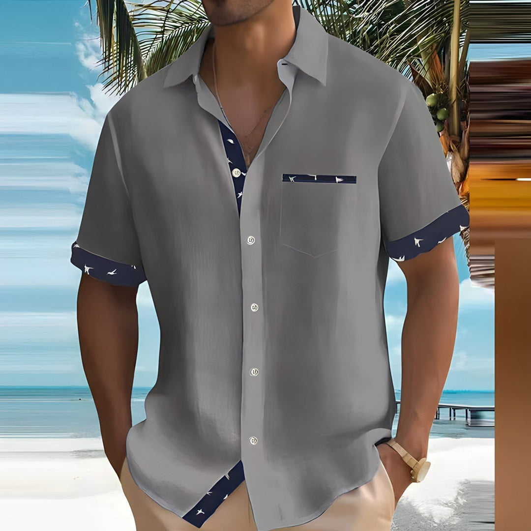 Homme en chemise grise à motifs, manches retroussées, sur une plage tropicale avec palmiers et mer turquoise en arrière-plan. Mode estivale décontractée.
