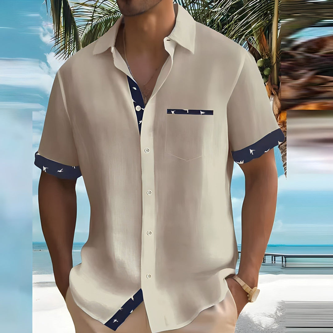 Homme en chemise beige à motifs marins, debout sur une plage tropicale avec palmiers et mer turquoise en arrière-plan, style estival décontracté.