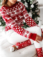 Femme en pull de Noël rouge et blanc avec motifs de rennes et flocons, assise devant un sapin décoré, portant des chaussettes assorties.