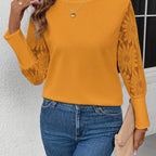 Femme portant un pull jaune à manches en dentelle florale, collier cœur, jean bleu, tenant une pochette beige devant un mur blanc.