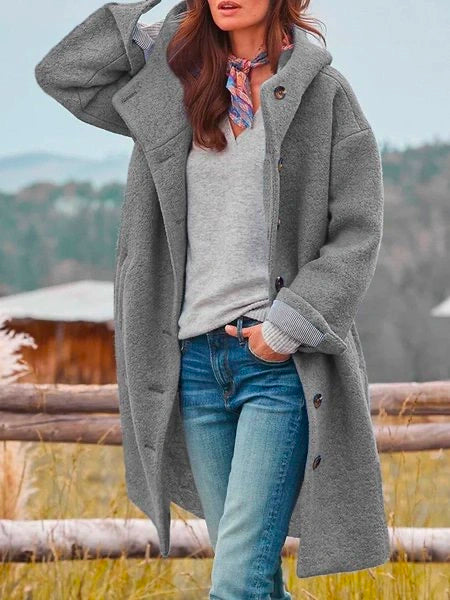 Femme portant un manteau gris oversize, pull gris, jeans bleu, foulard coloré, posant devant un paysage naturel. Mode automne-hiver tendance.