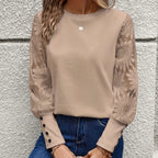 Femme portant un pull beige à manches longues en dentelle florale, boutons décoratifs, col rond, avec un collier, posant devant un mur texturé.