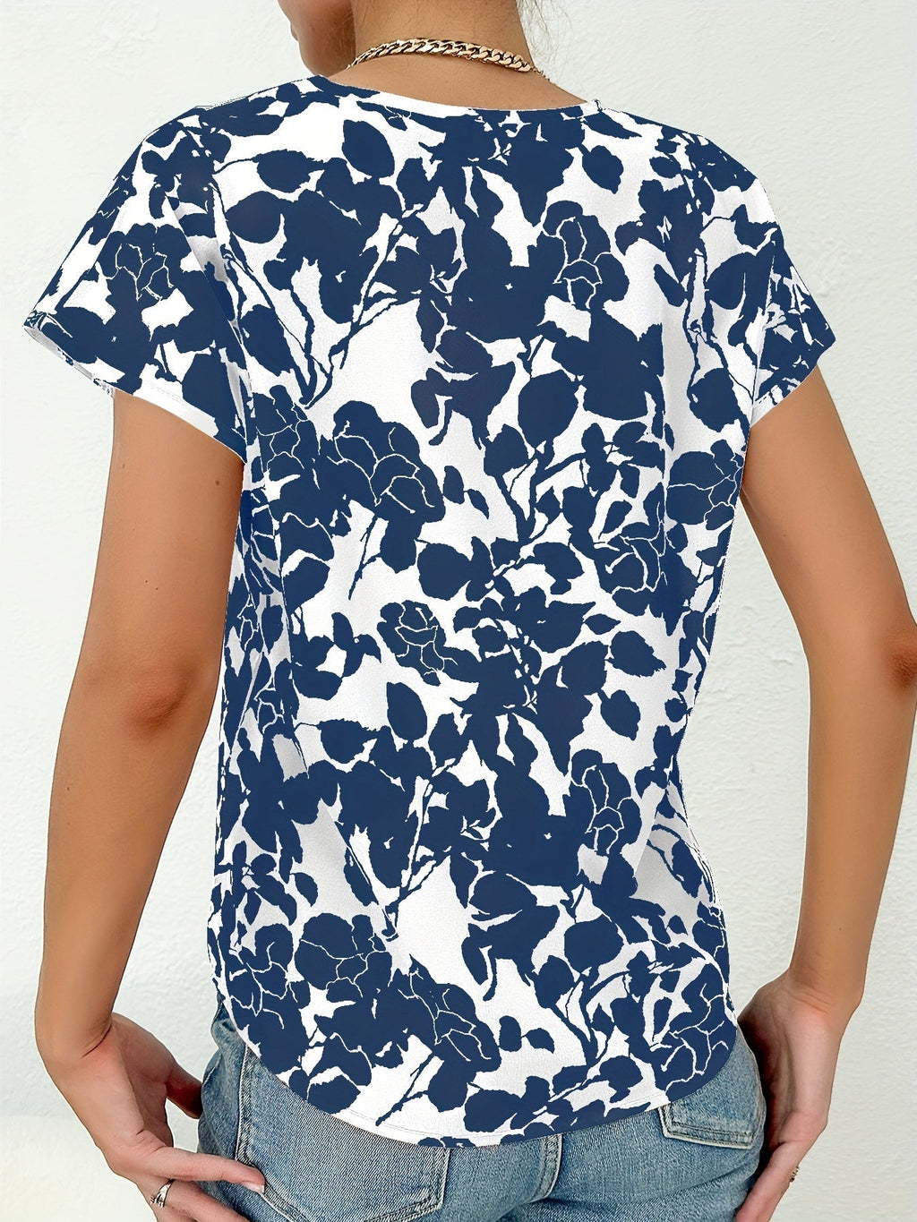 Femme portant un t-shirt à motifs floraux bleus et blancs, dos visible, style décontracté, chaîne argentée, fond blanc texturé.