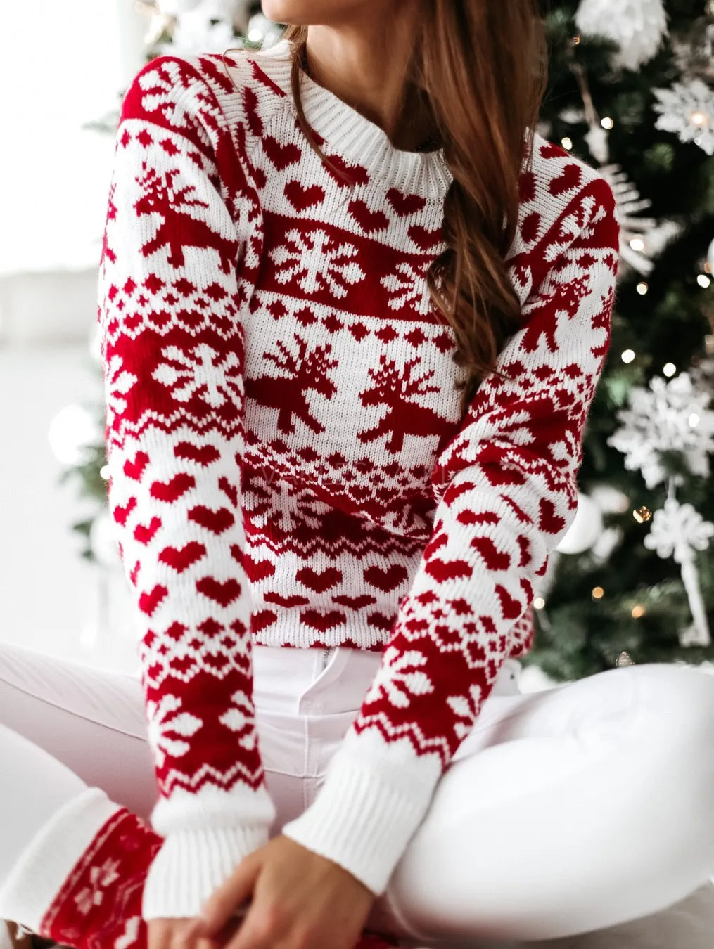 Femme portant un pull de Noël rouge et blanc avec motifs de rennes et flocons, assise devant un sapin décoré. Mode festive hiver.