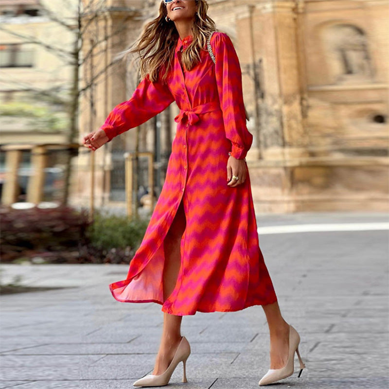 Femme souriante en robe longue rouge à motifs, marchant en talons hauts beiges dans une rue urbaine. Mode élégante, tenue chic, style moderne.