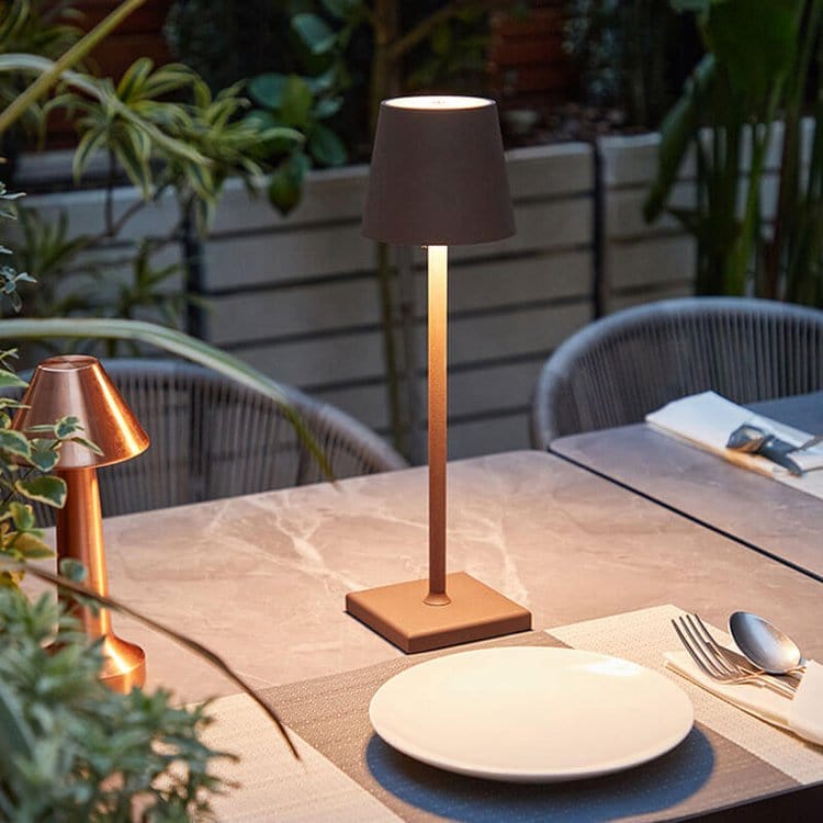 Lampe de table LED moderne sur une terrasse élégante, éclairant une table en marbre avec assiettes blanches, entourée de plantes vertes.