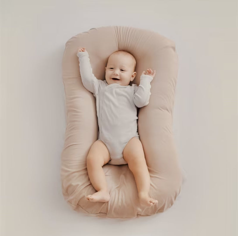 Bébé souriant allongé sur un coussin de soutien beige, vêtu d'un body blanc. Confort, sécurité, sommeil, accessoires bébé.