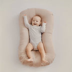 Bébé souriant allongé sur un coussin de soutien beige, vêtu d'un body blanc. Confort, sécurité, sommeil, accessoires bébé.