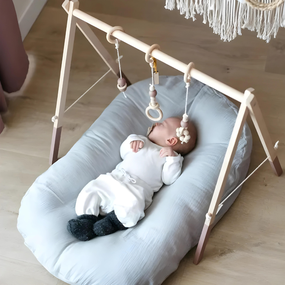 Bébé dormant sur un coussin gris sous un portique en bois avec jouets suspendus. Chambre d'enfant moderne, décoration scandinave, confort.