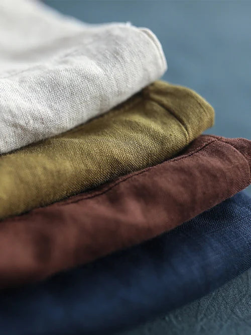 Quatre chemises en lin empilées, couleurs blanc, vert olive, marron et bleu marine, sur fond bleu. Mode durable, vêtements en lin.