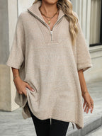 Femme portant un pull poncho beige à col zippé, manches courtes, accessoirisé de bijoux dorés, posant devant un bâtiment moderne. Mode automne chic.