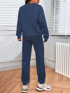 Personne portant un survêtement bleu marine, debout dans une pièce lumineuse avec parquet en bois, radiateur blanc et stores vénitiens.