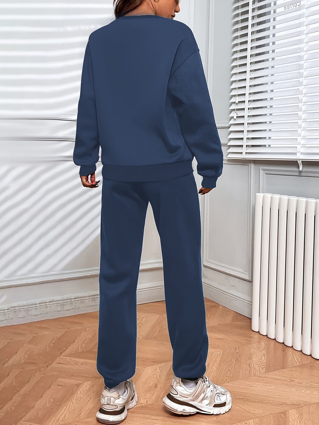 Personne portant un survêtement bleu marine, debout dans une pièce lumineuse avec parquet en bois, radiateur blanc et stores vénitiens.