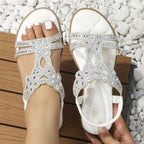 Sandales blanches ornées de strass, design floral, portées sur une terrasse en bois avec des galets blancs. Mode estivale élégante et tendance.