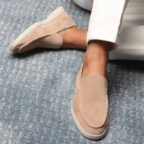 Mocassins en daim beige pour homme sur tapis gris, portés avec un pantalon blanc. Chaussures élégantes, confortables, style décontracté chic.