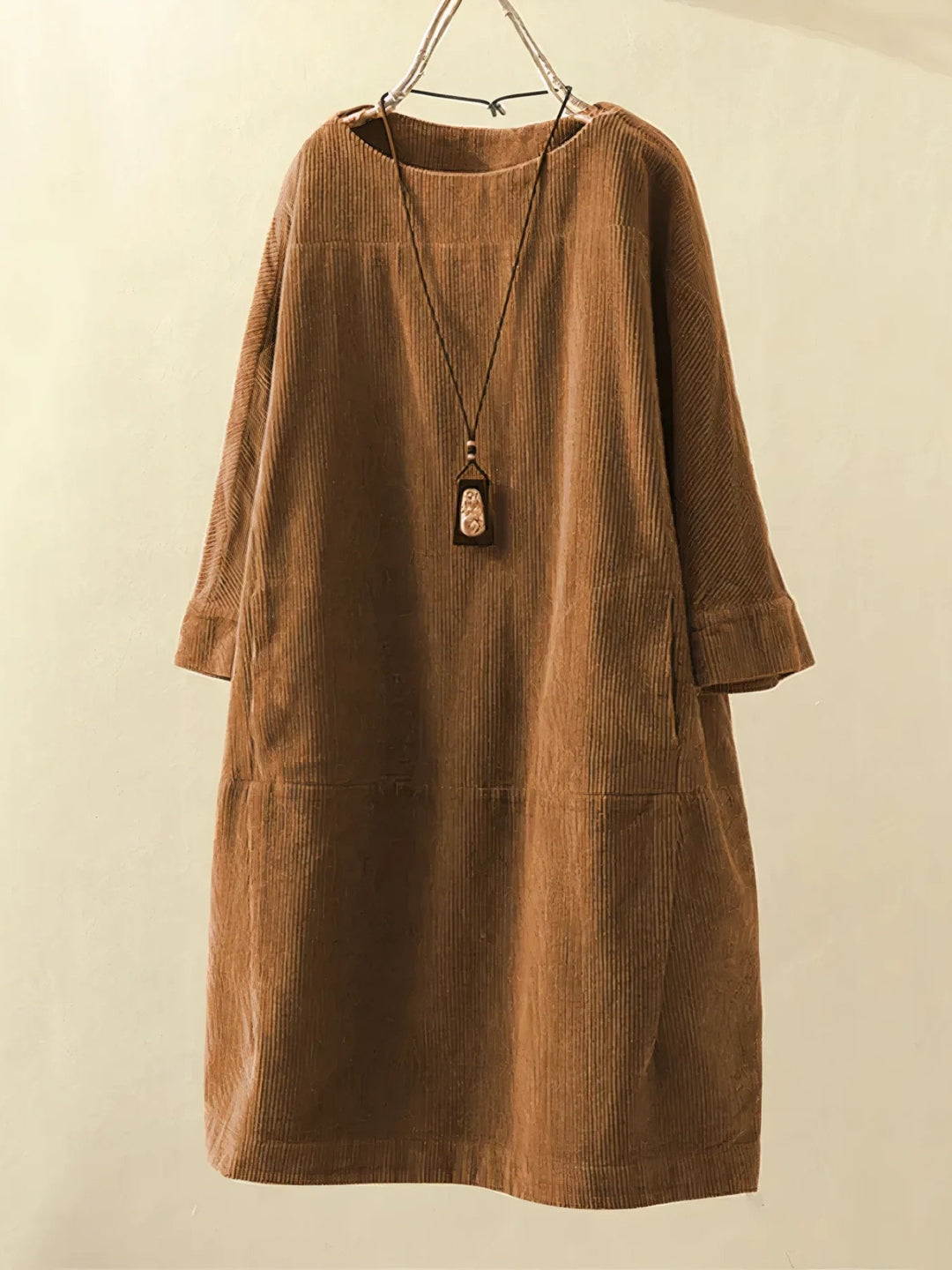 Robe en velours côtelé marron suspendue sur un cintre, avec un collier pendentif en bois. Mode automne, style décontracté, vêtement femme élégant.