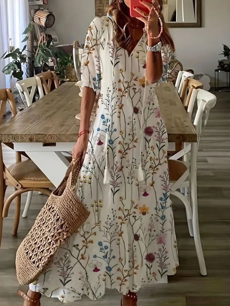 Robe longue bohème à motifs floraux colorés, femme tenant un sac en osier, intérieur moderne avec table en bois et chaises blanches.