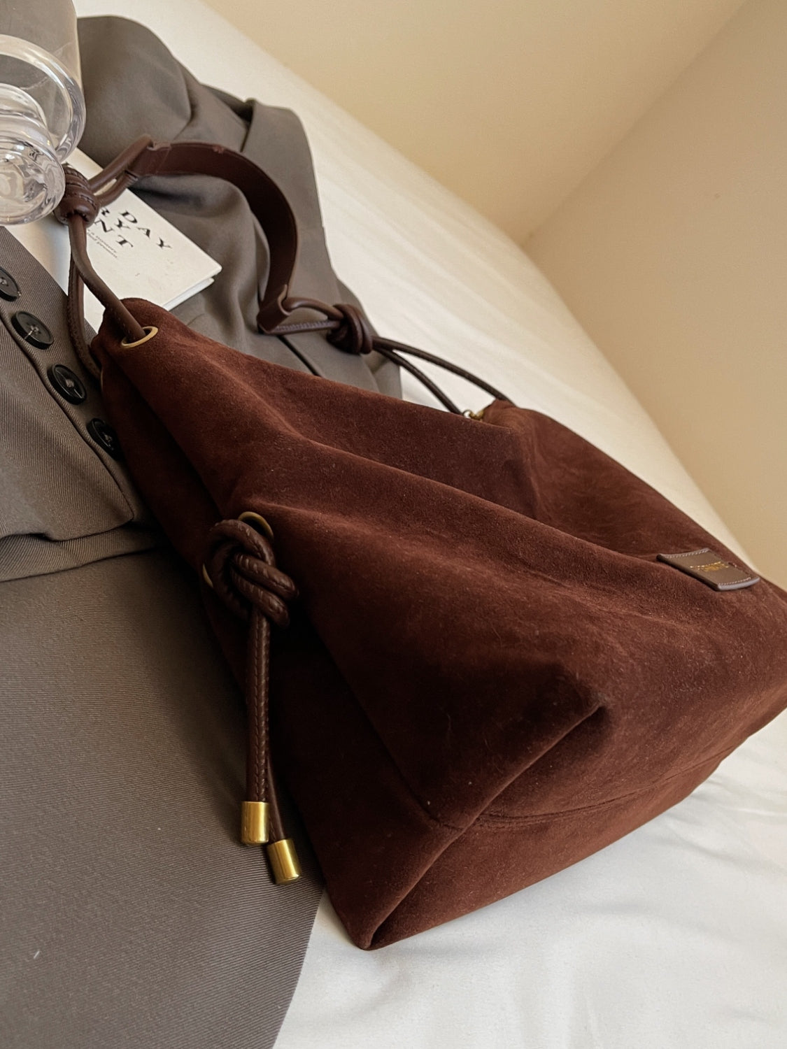 Sac à main en daim marron sur un manteau gris, avec des poignées en cuir et des détails dorés, posé sur un lit blanc. Accessoire mode élégant.