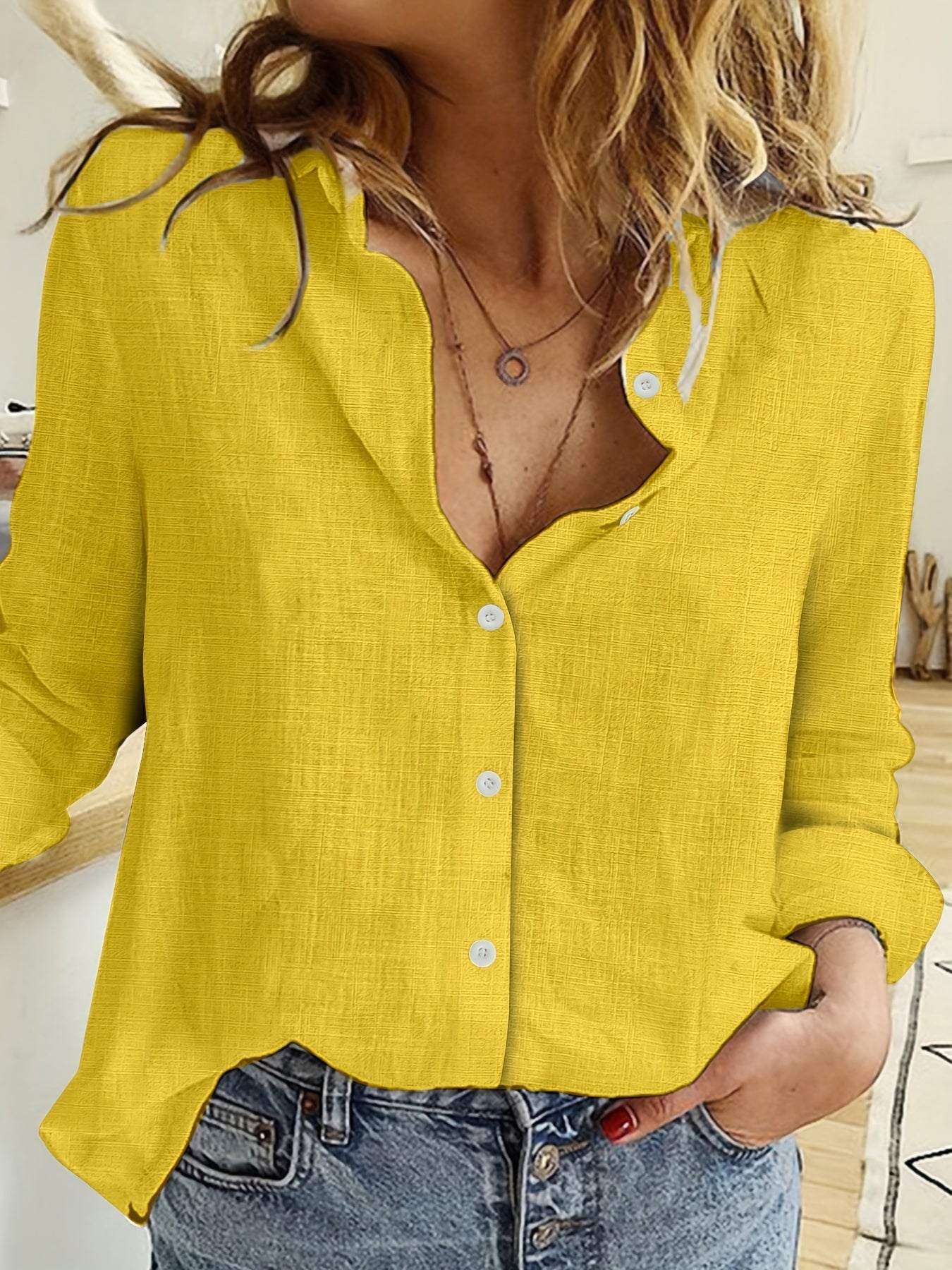 Femme portant une chemise jaune en lin, manches retroussées, col ouvert, avec un collier délicat, posant dans un intérieur moderne et lumineux.