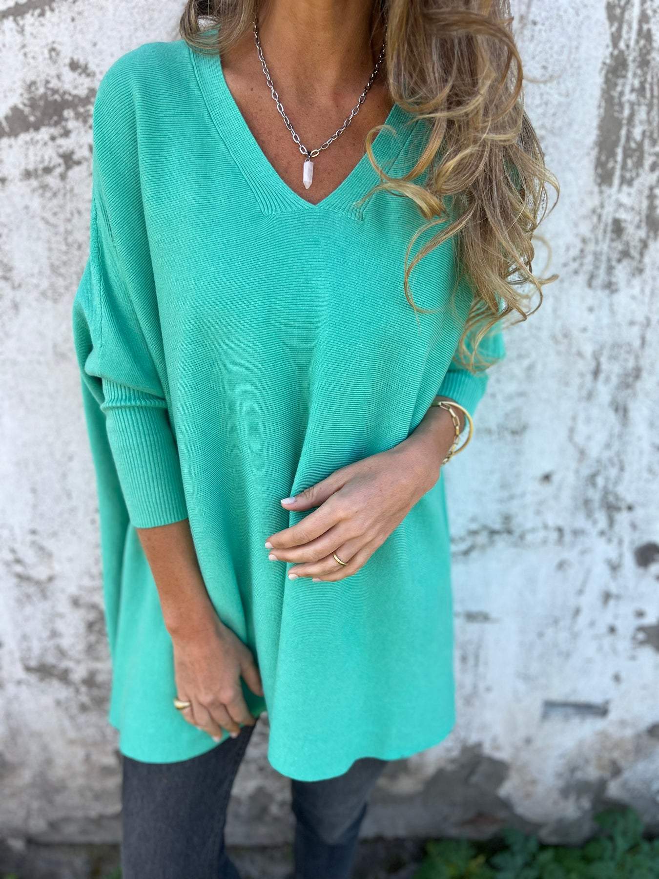 Femme portant un pull vert menthe ample, col en V, manches longues, devant un mur texturé. Mode décontractée, tendance automne-hiver.