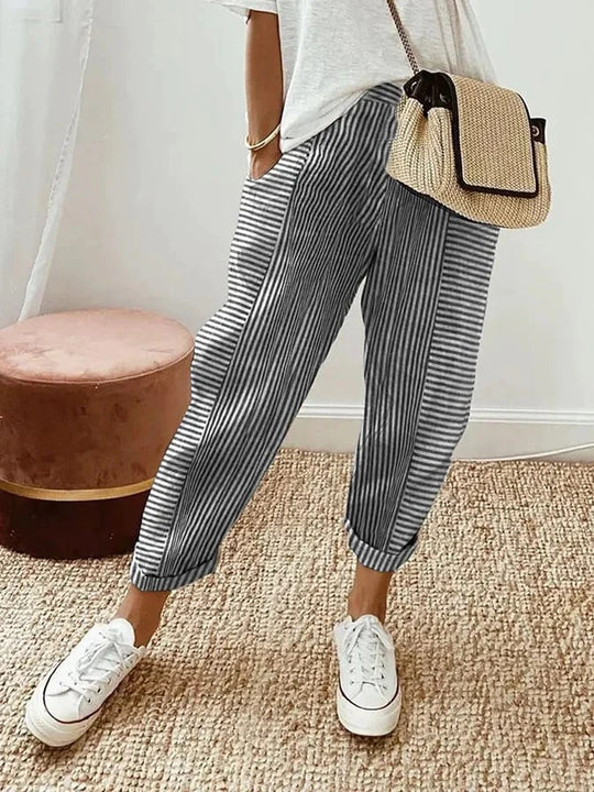 Pantalon rayé noir et blanc, style décontracté, porté avec un t-shirt blanc et des baskets blanches, sac en osier, sur tapis beige.