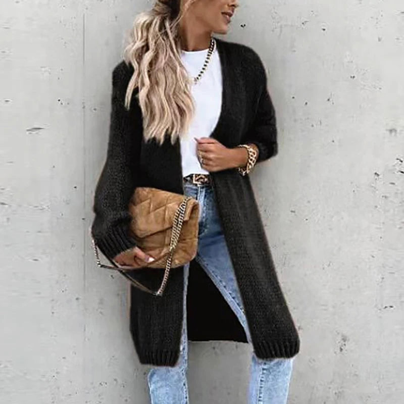 Femme portant un long cardigan noir, t-shirt blanc, jean bleu, sac à main marron, posant contre un mur gris. Mode automne chic et décontractée.