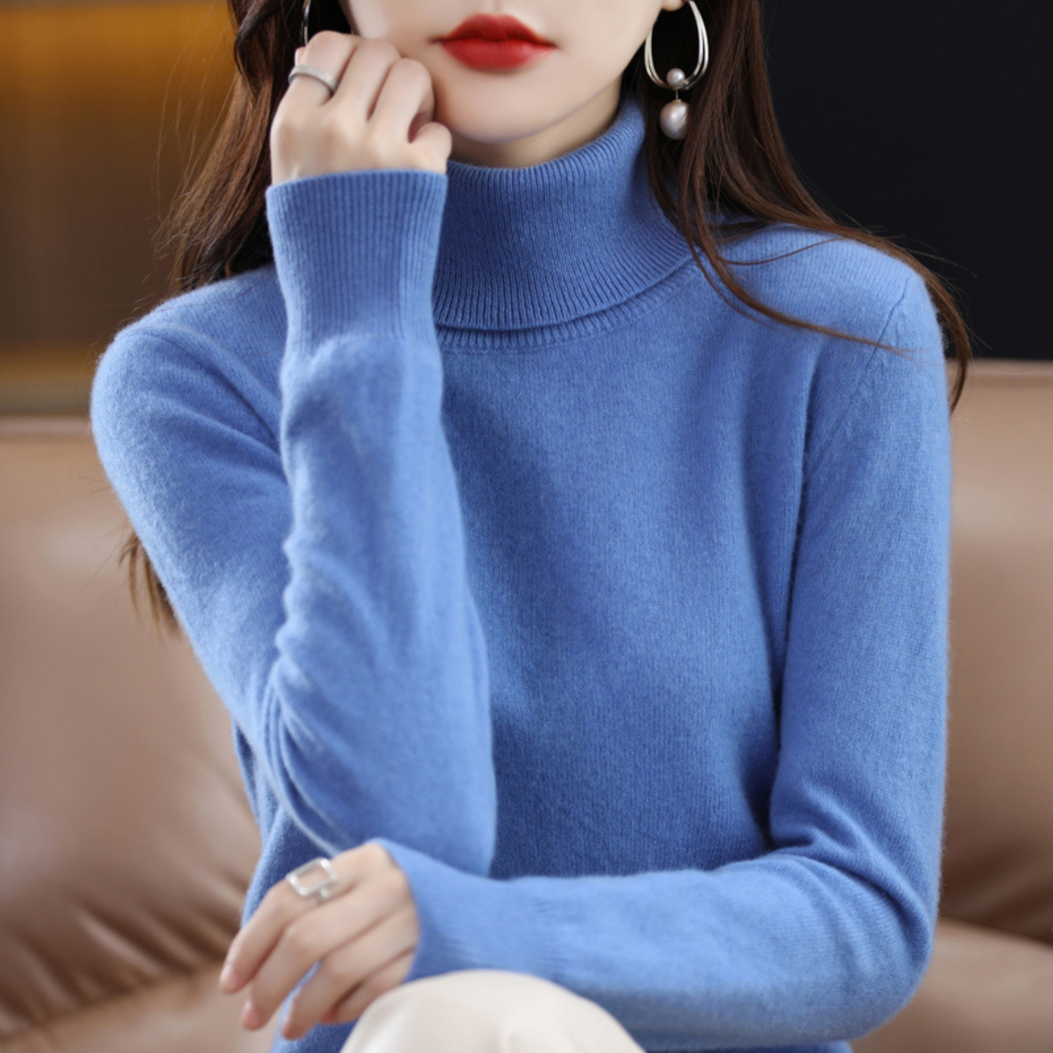 Femme portant un pull bleu col roulé, assise sur un canapé en cuir. Mode automne-hiver, style élégant, accessoires bijoux argentés.