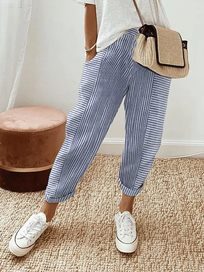 Femme portant un pantalon rayé bleu et blanc, t-shirt blanc, baskets blanches et sac en osier. Mode décontractée, tendance estivale.