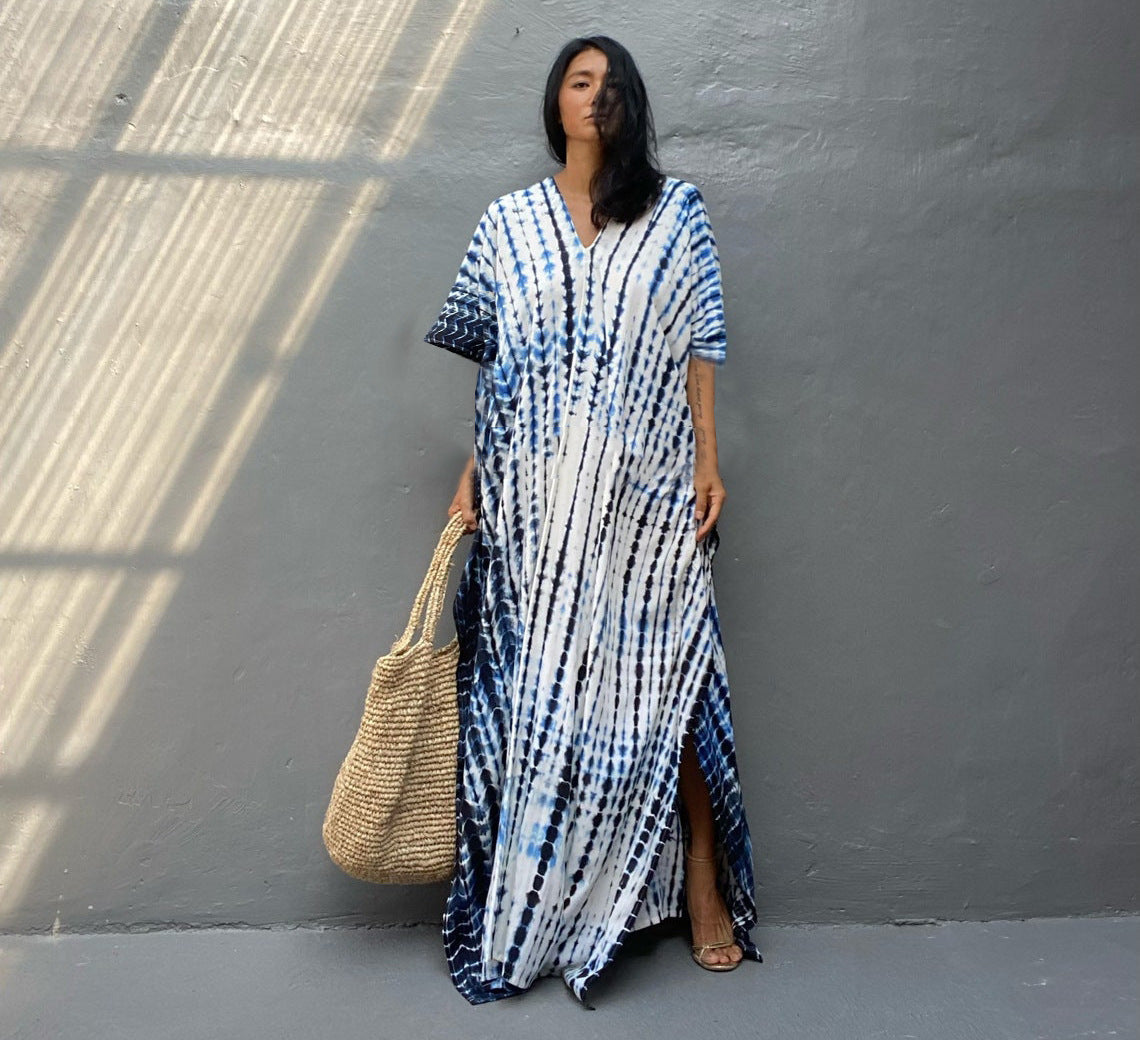 Femme portant une robe longue tie-dye bleue et blanche, tenant un grand sac en osier, posant contre un mur gris. Mode estivale, style décontracté.
