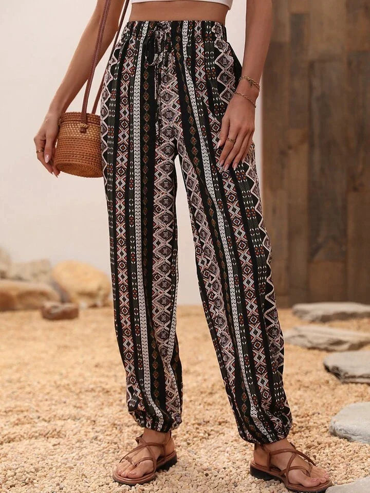 Pantalon ample à motifs ethniques noirs et blancs, porté avec un sac en osier et des sandales marron, sur fond de sol en pierre et bois.