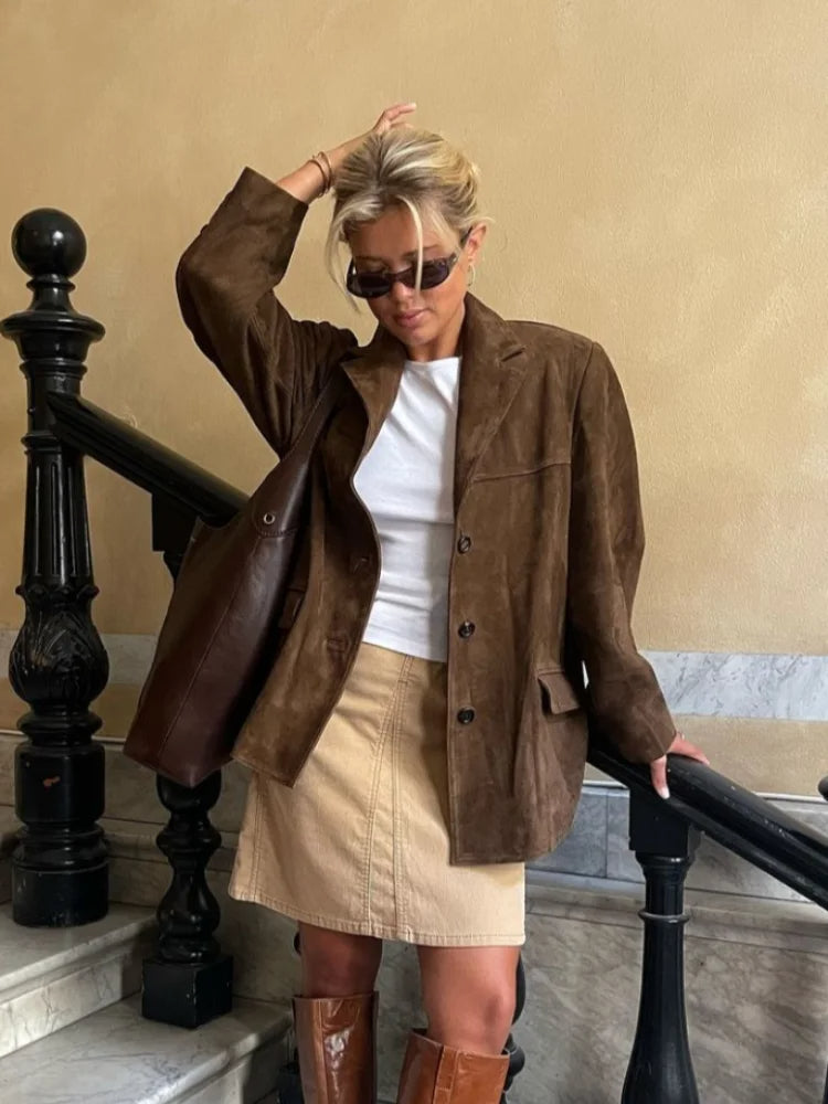Femme en veste marron, jupe beige et bottes en cuir, posant sur un escalier intérieur. Style automnal chic, lunettes de soleil, sac en cuir.