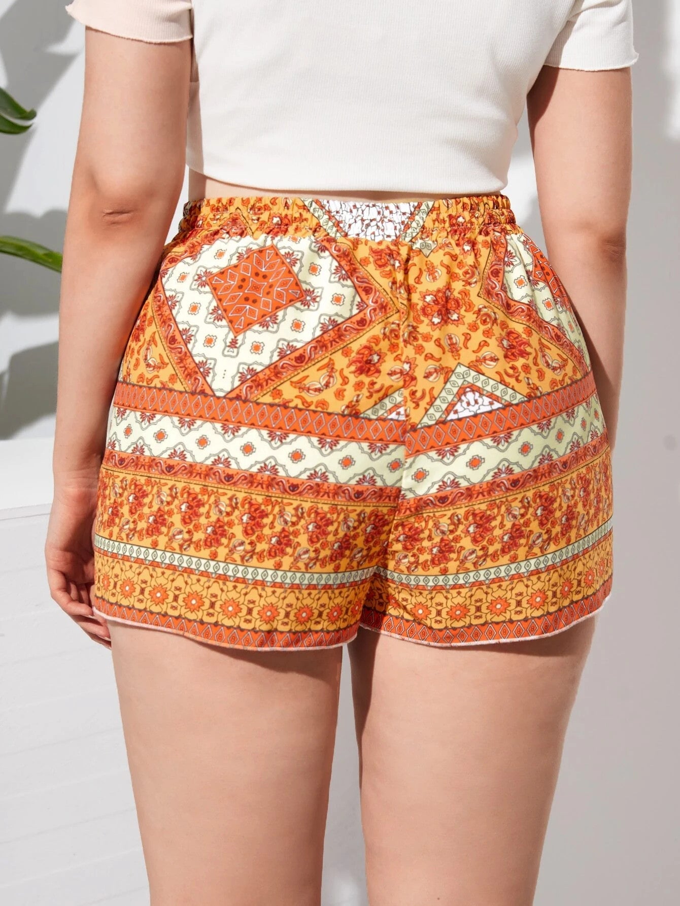 Femme portant un short à motifs bohèmes orange et blanc, style estival, fond blanc, mode tendance, tenue décontractée, vue de dos.