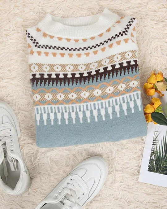 Pull-over en laine à motifs géométriques colorés, posé sur un tapis beige, entouré de baskets blanches et d'un magazine avec des fleurs jaunes.