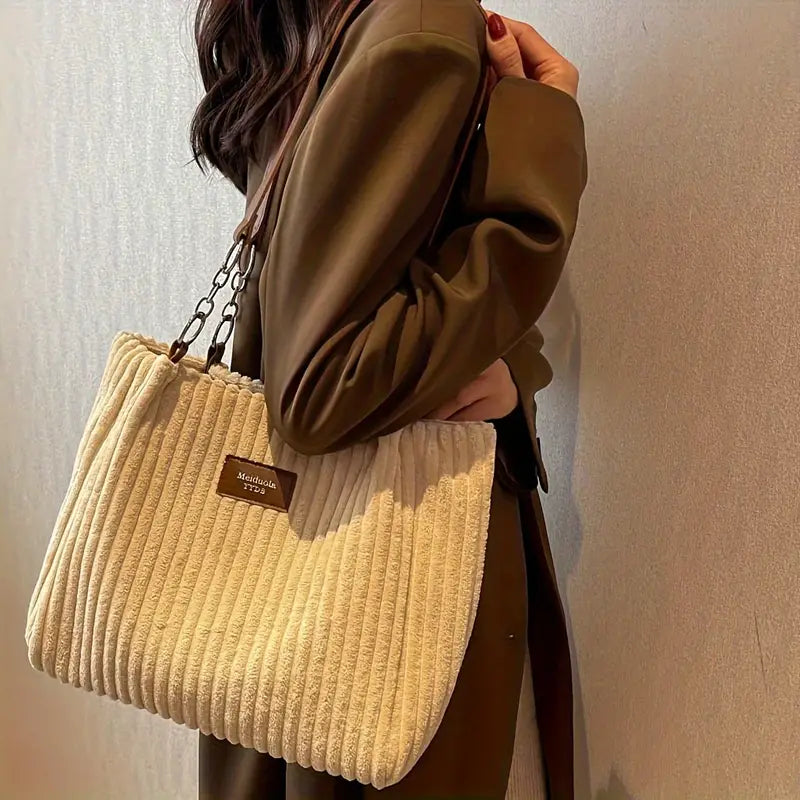 Sac à main en velours côtelé beige porté par une femme en manteau marron, avec une étiquette en cuir marron. Accessoire mode élégant et tendance.