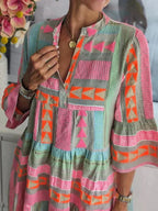 Robe bohème colorée avec motifs géométriques roses, oranges et verts, manches évasées, col en V, femme portant des bijoux dorés. Mode estivale tendance.