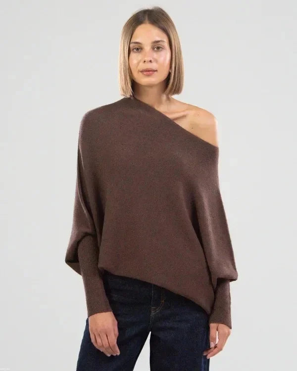 Femme portant un pull asymétrique marron, épaule dénudée, avec un jean bleu. Mode automne-hiver, style décontracté et élégant.